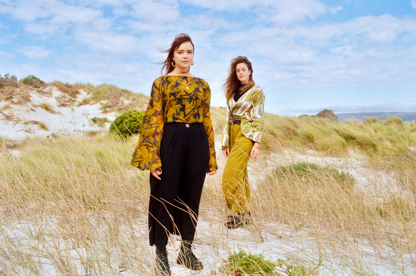 Two women standing on a grassy hill with a blue sky and ocean in the background