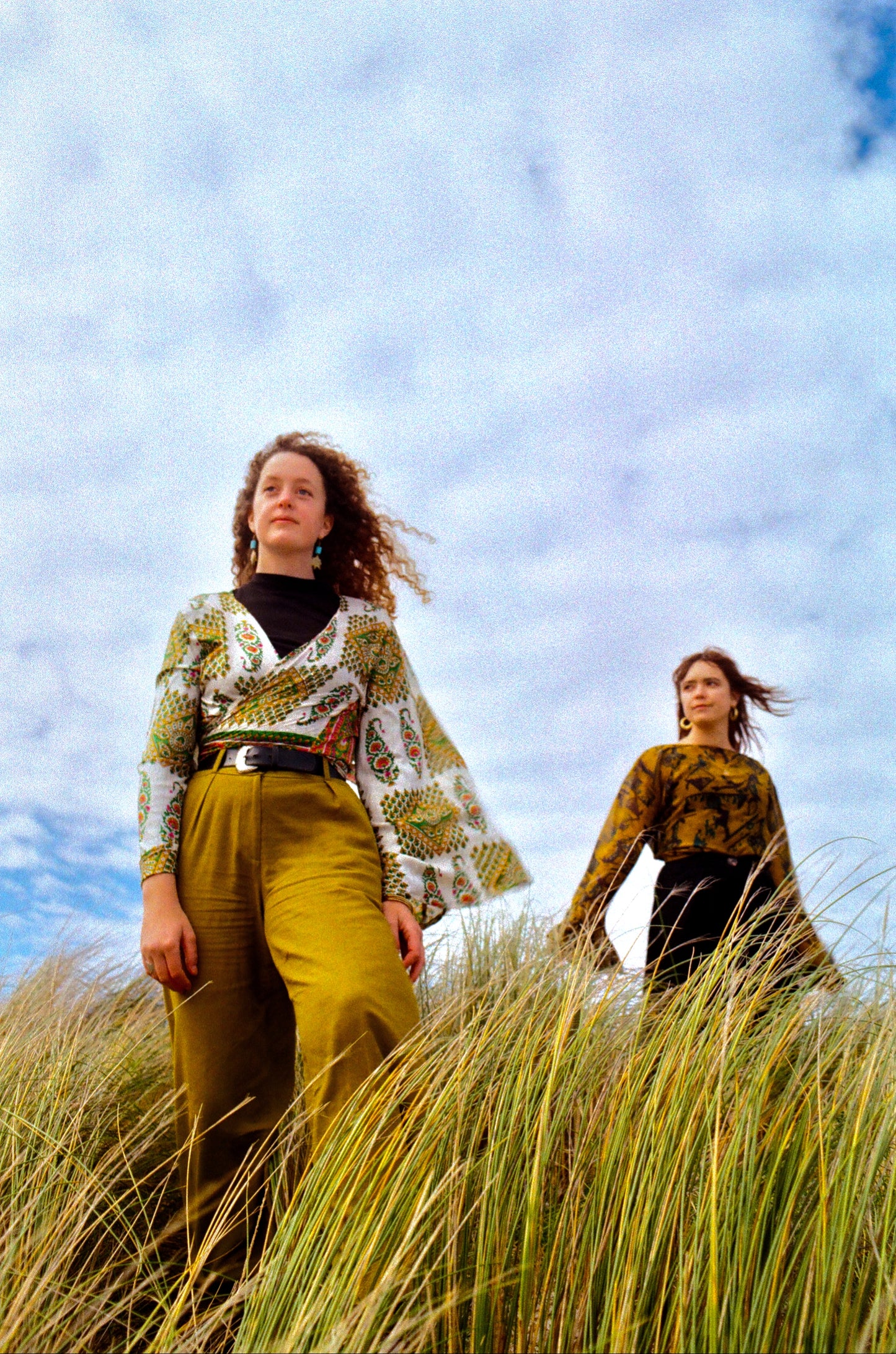 Two women standing in tall grass with a blue sky and clouds in the background