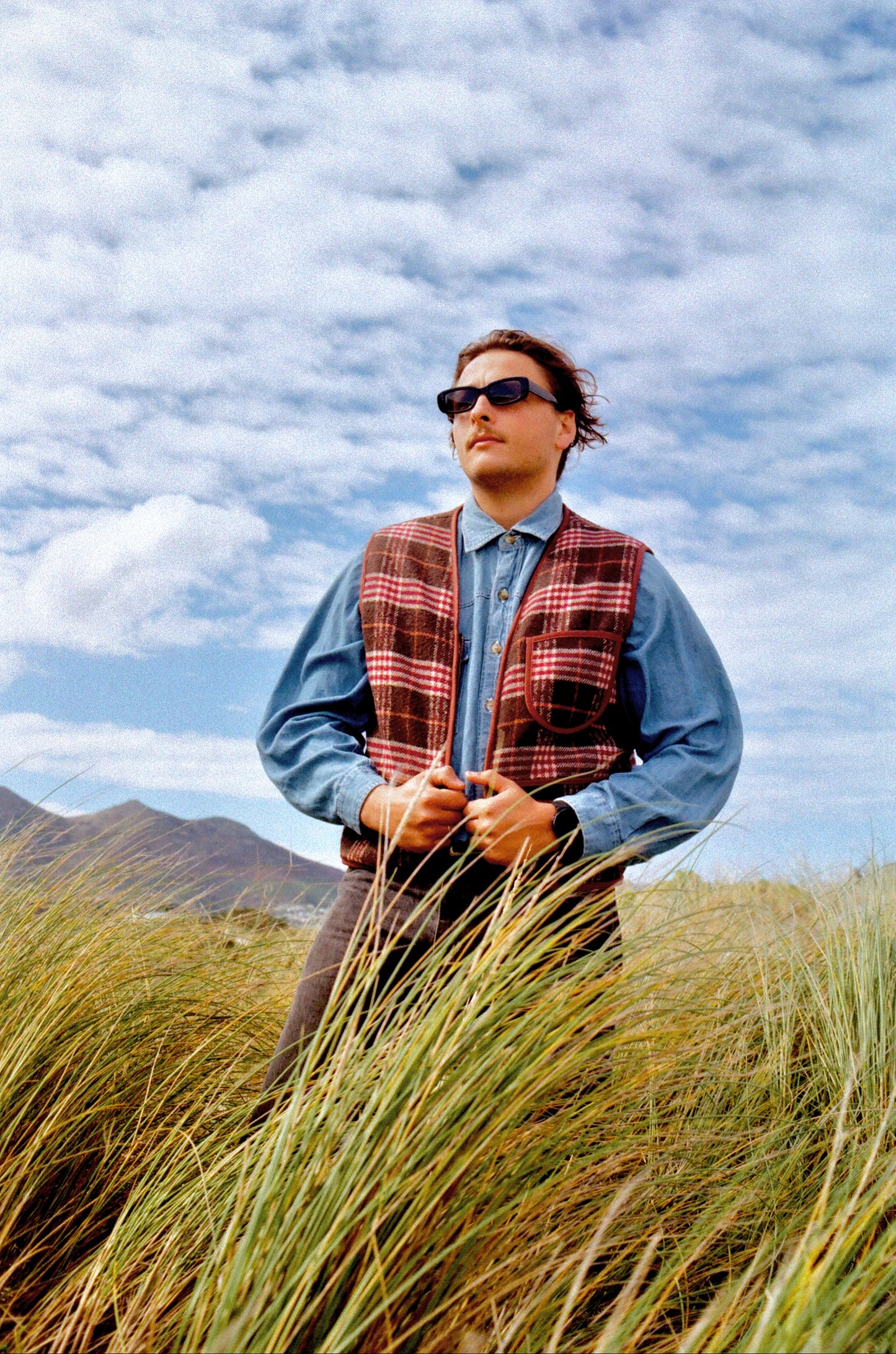 Man wearing sunglasses and a plaid vest standing in tall grass with a mountainous background vintage look