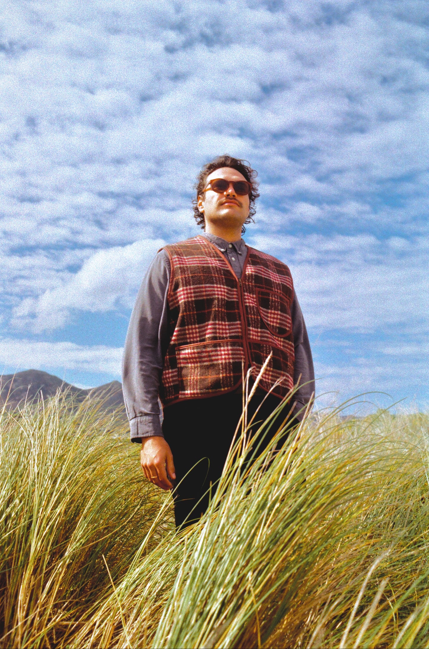 Man wearing a plaid vest standing in tall grass with a blue sky and clouds in the background