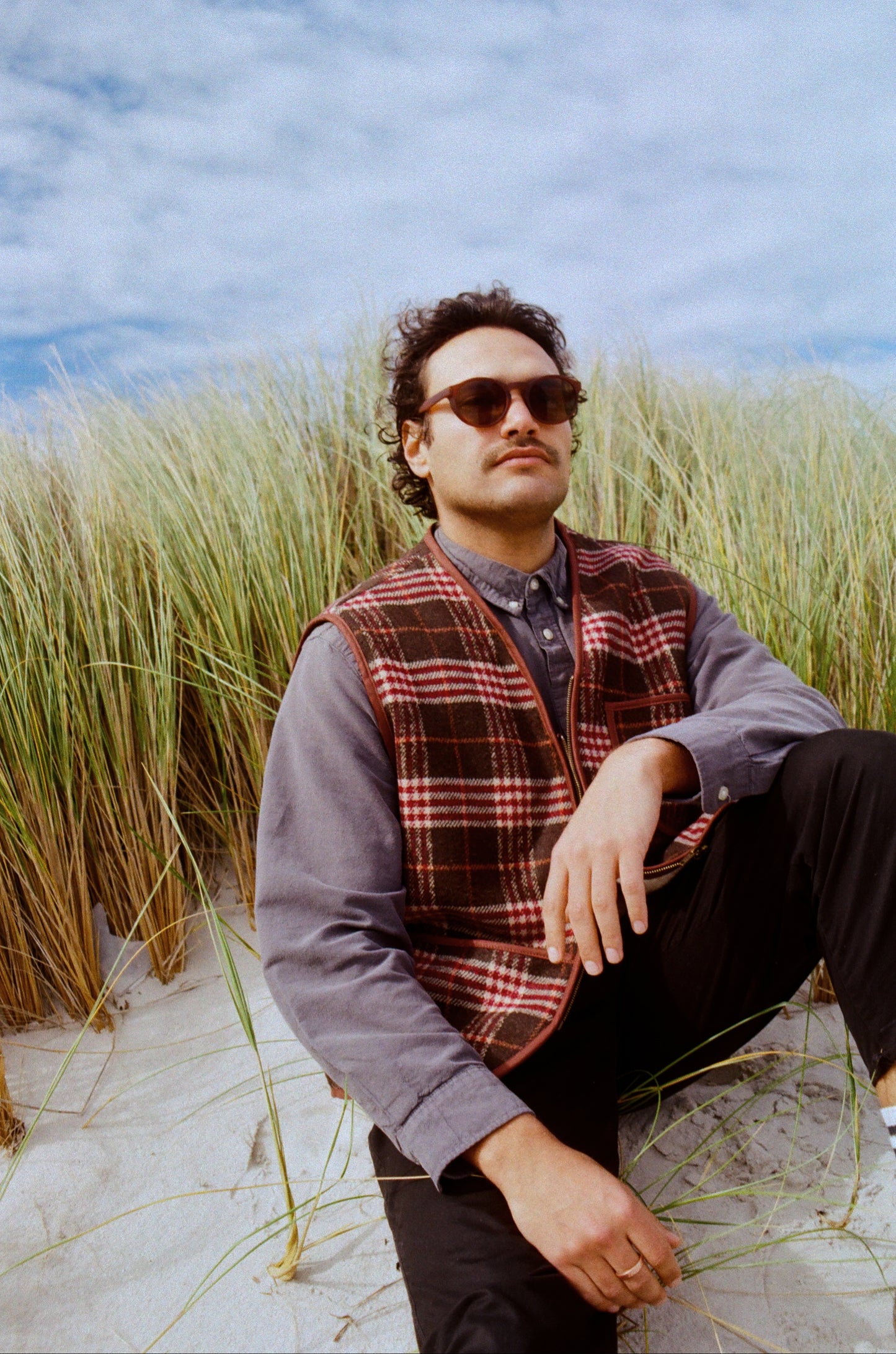 Man wearing sunglasses and a plaid vest sitting in tall grass with a blue sky background
