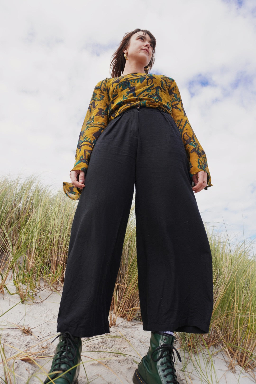 Person wearing a yellow patterned top and black pants standing on a sandy beach with grass.