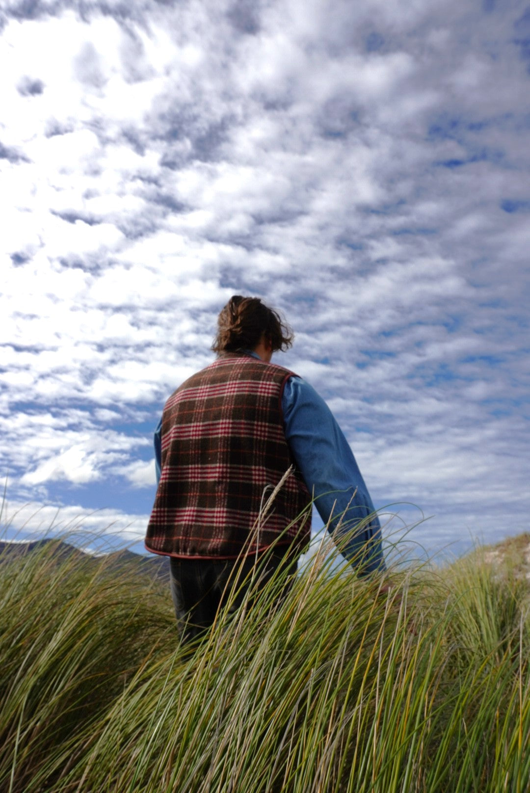Person standing in tall grass looking up at a cloudy sky wearing a plaid vest