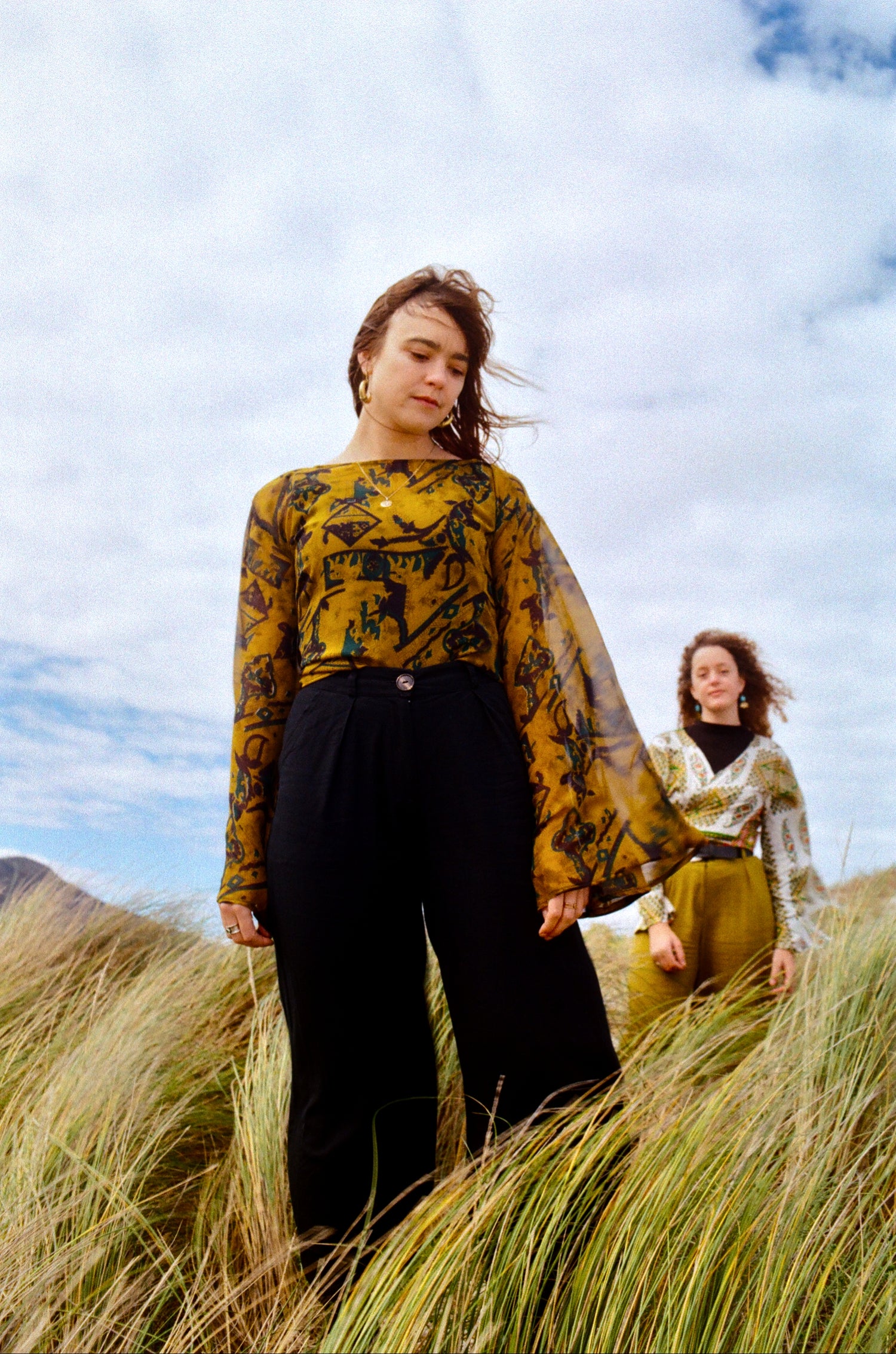 Two people standing in a grassy field with a blue sky wearing high waisted pants