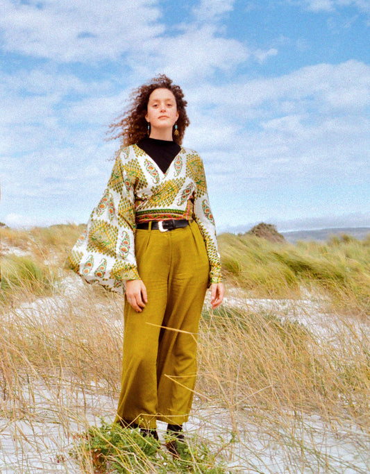 Woman in a patterned top and green pants standing on a grassy hill with a blue sky.